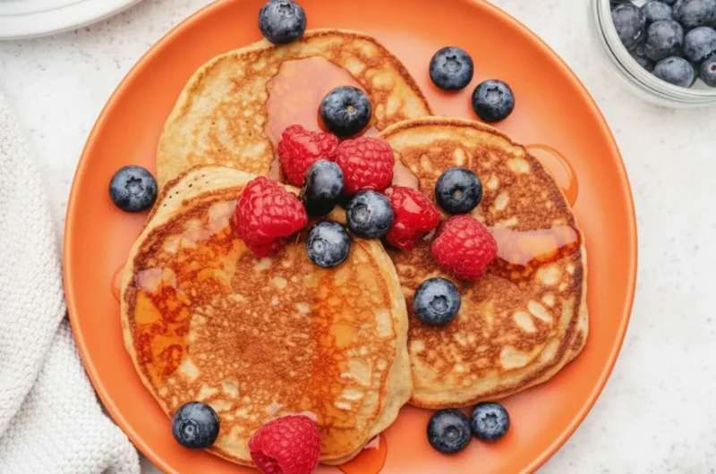 A hero shot of three fluffy Cottage Cheese Pancakes stacked on a minimalist white ceramic plate. The pancakes are golden-brown with slightly uneven, rustic edges. A generous pile of fresh red raspberries and dark blue blueberries sits on top, with amber maple syrup drizzled over the fruit and pooling slightly on the plate. The scene is set on a white marble countertop with soft morning light coming from the side, creating gentle shadows. A wooden cutting board is partially visible in the corner.