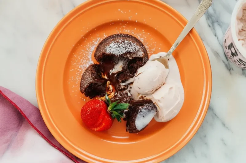A hero shot of a Mini Lava Cake for Two on a white scalloped ceramic plate, the cake is broken open with thick chocolate lava flowing out, dusted with powdered sugar, a scoop of pale pink strawberry ice cream on the side, a whole fresh red strawberry with green leaves, vintage silver spoon resting on the plate, marble countertop background, soft morning light from the side, warm tones, 4:3 aspect ratio.