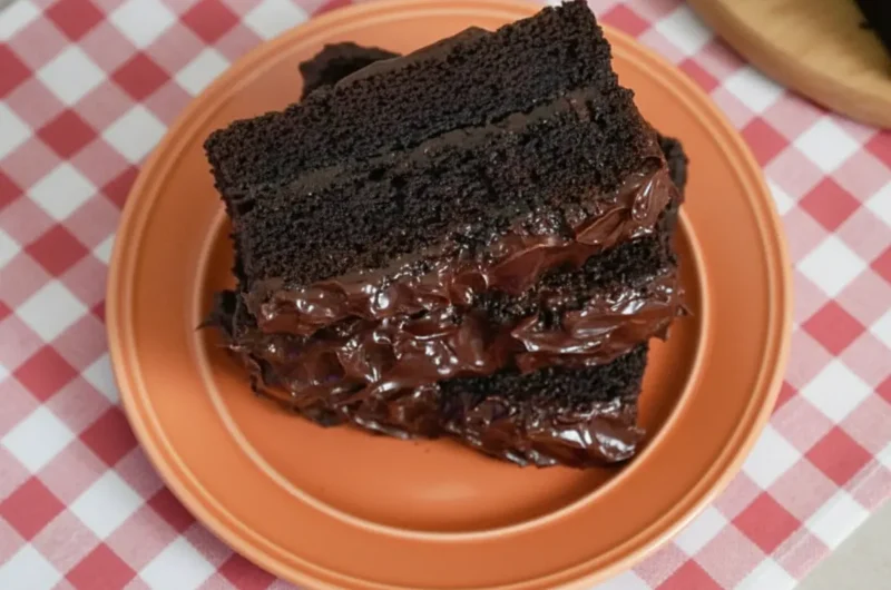 A stack of three thick, dense slices of moist chocolate banana cake on a minimalist white plate, set on a marble countertop with soft morning light from an east window. The cake slices have a rich, dark fudgy crumb and are topped with thick, glossy chocolate frosting showing beautiful swoops and peaks. The background features a rustic wood cutting board and soft shadows, capturing a warm, inviting kitchen scene.