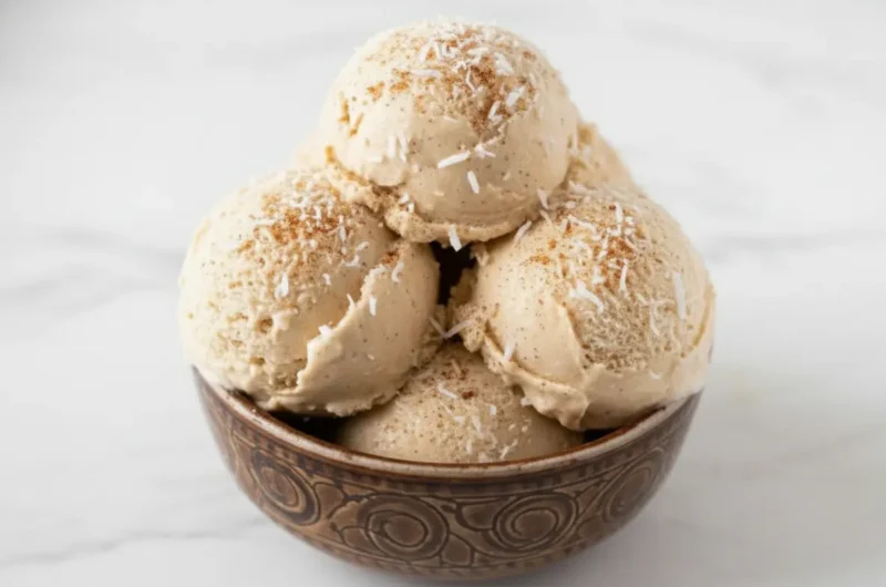 A wide 4:3 shot on a marble countertop in bright morning light. A brown ceramic bowl with a carved spiral pattern sits on a weathered wooden cutting board. Inside the bowl are three generous, textured scoops of beige No-Churn Coquito Ice Cream, showing a creamy, slightly granular texture. In the soft-focus background, a whole brown coconut and lush green leaves are visible near a clean white wall. Soft shadows and warm tones create a cozy, lived-in kitchen atmosphere.
