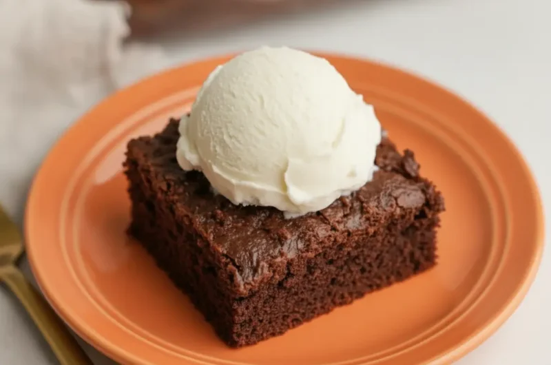 A square slice of dark Chocolate Crunch Cake served on a minimalist white ceramic plate. The cake has a thick, jagged topping of chocolate-coated crispy cereal. On top of the crunch layer is a large, perfectly round scoop of vanilla ice cream that is starting to melt and drip down the sides. The plate sits on a white marble countertop with a soft wooden accent nearby. The background is softly blurred, showing the rest of the cake in a glass baking dish. High-end food photography, natural morning light from an east window, warm tones, 4:3 aspect ratio, no hands, no people.