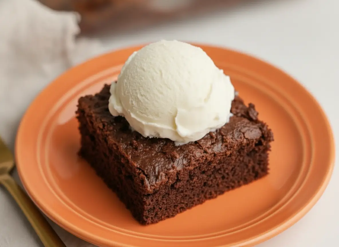 A square slice of dark Chocolate Crunch Cake served on a minimalist white ceramic plate. The cake has a thick, jagged topping of chocolate-coated crispy cereal. On top of the crunch layer is a large, perfectly round scoop of vanilla ice cream that is starting to melt and drip down the sides. The plate sits on a white marble countertop with a soft wooden accent nearby. The background is softly blurred, showing the rest of the cake in a glass baking dish. High-end food photography, natural morning light from an east window, warm tones, 4:3 aspect ratio, no hands, no people.