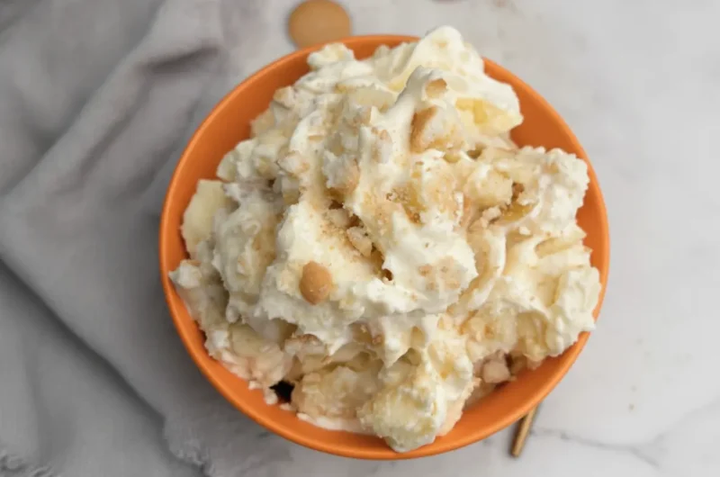 A high-angle 4:3 hero shot of a large white ceramic bowl filled with Pound Cake Banana Pudding. The pudding is creamy yellow and white with visible golden-brown cubes of pound cake and round vanilla wafers. The top is peaked with fluffy whipped cream and sprinkled with cake crumbs. The bowl sits on a white marble countertop next to a dark wooden cutting board. Soft morning light enters from the side, creating gentle shadows. Fresh mint is tucked in the background.