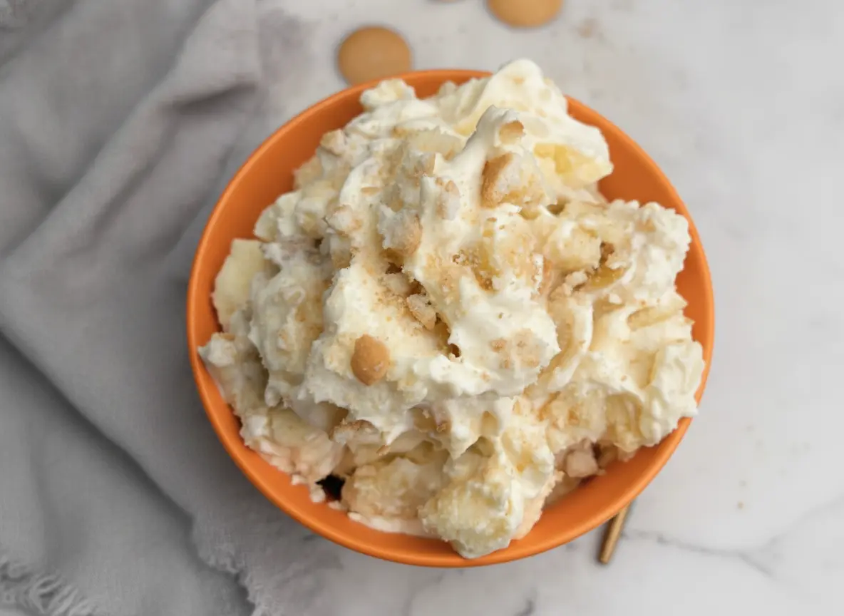 A high-angle 4:3 hero shot of a large white ceramic bowl filled with Pound Cake Banana Pudding. The pudding is creamy yellow and white with visible golden-brown cubes of pound cake and round vanilla wafers. The top is peaked with fluffy whipped cream and sprinkled with cake crumbs. The bowl sits on a white marble countertop next to a dark wooden cutting board. Soft morning light enters from the side, creating gentle shadows. Fresh mint is tucked in the background.