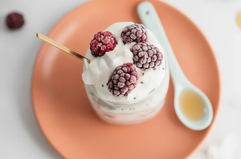 A hero shot of a glass mason jar filled with Protein Overnight Oats With Coconut Milk, featuring visible purple and red swirls from frozen berries. The jar is topped with frosty blackberries and white coconut flakes. A thin wooden spoon is inserted at an angle. The setting is a bright kitchen with marble countertops, wood accents, and soft morning light from an east-facing window. 4:3 aspect ratio.
