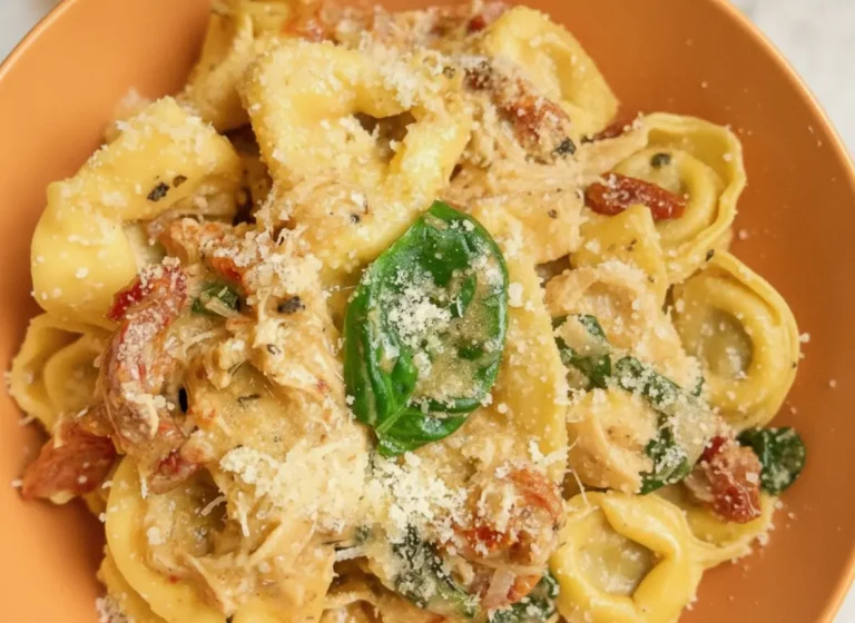 A high-angle hero shot of a white bowl filled with Slow Cooker Marry Me Tortellini. The pasta is cheese-filled tortellini coated in a thick, creamy orange-tinted sauce with visible bits of red sun-dried tomatoes and wilted green spinach. A gold fork rests in the bowl, lifting a single tortellini. The bowl sits on a white marble countertop next to a wooden cutting board with fresh basil leaves. Natural morning light creates soft shadows and warm tones.