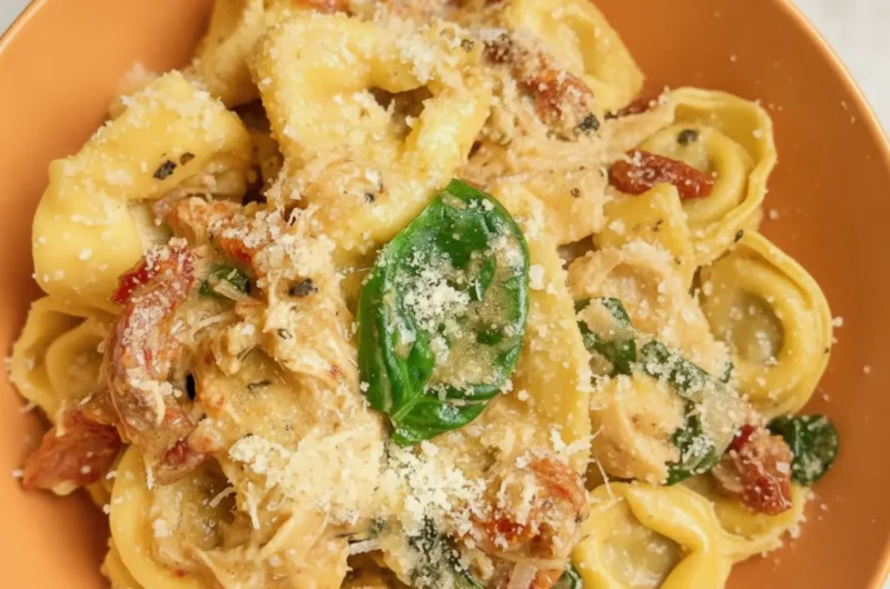 A high-angle hero shot of a white bowl filled with Slow Cooker Marry Me Tortellini. The pasta is cheese-filled tortellini coated in a thick, creamy orange-tinted sauce with visible bits of red sun-dried tomatoes and wilted green spinach. A gold fork rests in the bowl, lifting a single tortellini. The bowl sits on a white marble countertop next to a wooden cutting board with fresh basil leaves. Natural morning light creates soft shadows and warm tones.