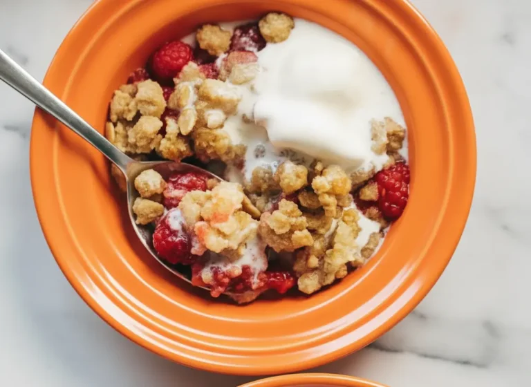 A high-angle 4:3 shot of two white ceramic ramekins filled with Two Person Raspberry Crumbles on a white marble countertop. The crumbles feature vibrant red bubbling raspberries and a golden-brown toasted oat topping. A generous dollop of white yogurt is melting over the top of one ramekin. A silver spoon is resting inside the other, showing the juicy interior. Soft morning light from an east window creates gentle shadows. Warm wood accents and fresh mint are blurred in the background.
