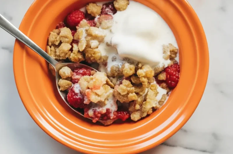 A high-angle 4:3 shot of two white ceramic ramekins filled with Two Person Raspberry Crumbles on a white marble countertop. The crumbles feature vibrant red bubbling raspberries and a golden-brown toasted oat topping. A generous dollop of white yogurt is melting over the top of one ramekin. A silver spoon is resting inside the other, showing the juicy interior. Soft morning light from an east window creates gentle shadows. Warm wood accents and fresh mint are blurred in the background.