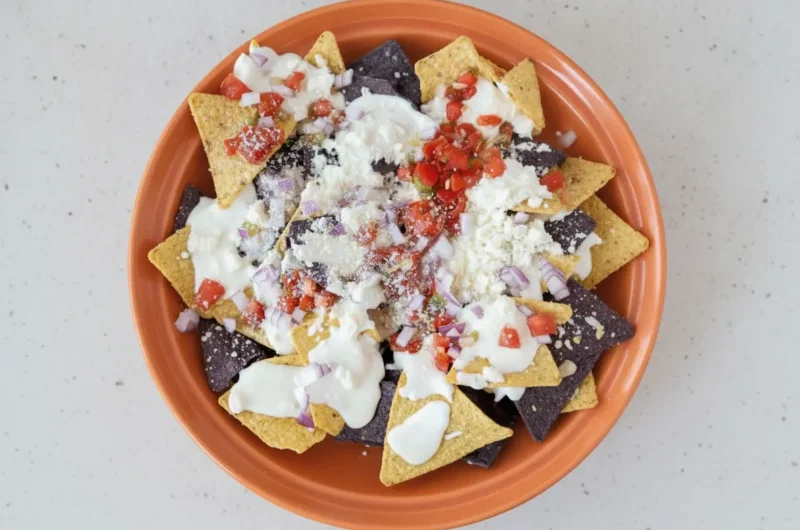 A high-angle 4:3 hero shot of Queso Blanco Nachos on a large white ceramic platter. The nachos consist of a mix of yellow and purple corn tortilla chips, generously drizzled with a thick, glossy white cheese sauce. The chips are topped with a heavy dusting of white crumbly cotija cheese, finely diced red onions, and bright red diced tomatoes. The platter sits on a white marble countertop with a hint of a wooden cutting board nearby. Soft morning light from an east window creates gentle shadows and highlights the creamy texture of the sauce.