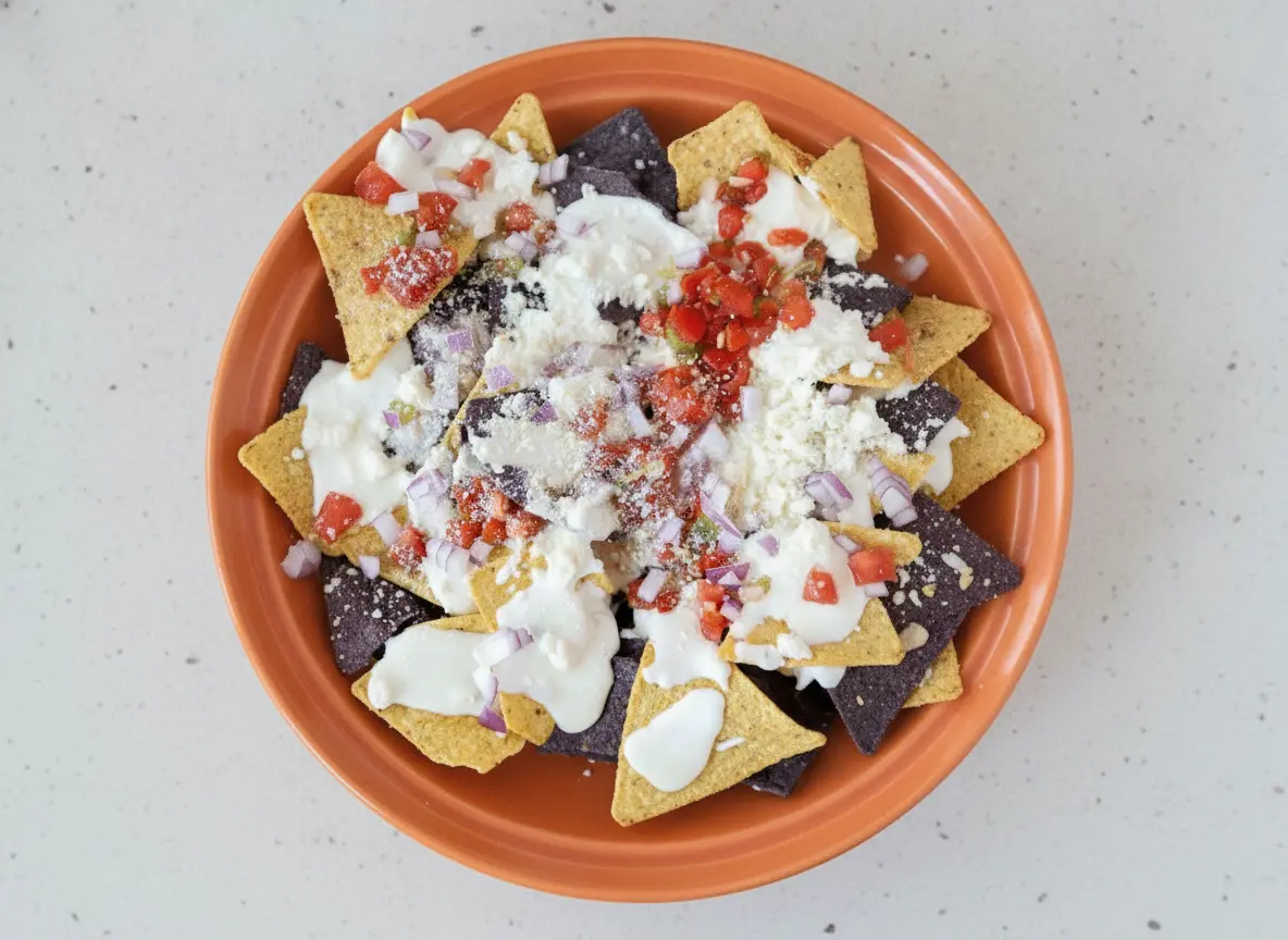 A high-angle 4:3 hero shot of Queso Blanco Nachos on a large white ceramic platter. The nachos consist of a mix of yellow and purple corn tortilla chips, generously drizzled with a thick, glossy white cheese sauce. The chips are topped with a heavy dusting of white crumbly cotija cheese, finely diced red onions, and bright red diced tomatoes. The platter sits on a white marble countertop with a hint of a wooden cutting board nearby. Soft morning light from an east window creates gentle shadows and highlights the creamy texture of the sauce.