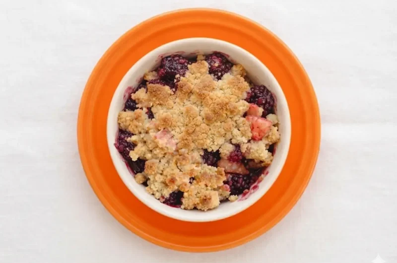 A hero shot of a rustic ceramic dish filled with Apple and Blackberry Crumble. The topping is golden-brown, buttery, and craggy with visible oats and sugar crystals. A large spoonful has been taken out, revealing the steaming, dark purple blackberry juices and bright red-stained apple chunks inside. The dish is set on a marble countertop with soft morning light from a side window, warm tones, and soft shadows. No hands, no people.