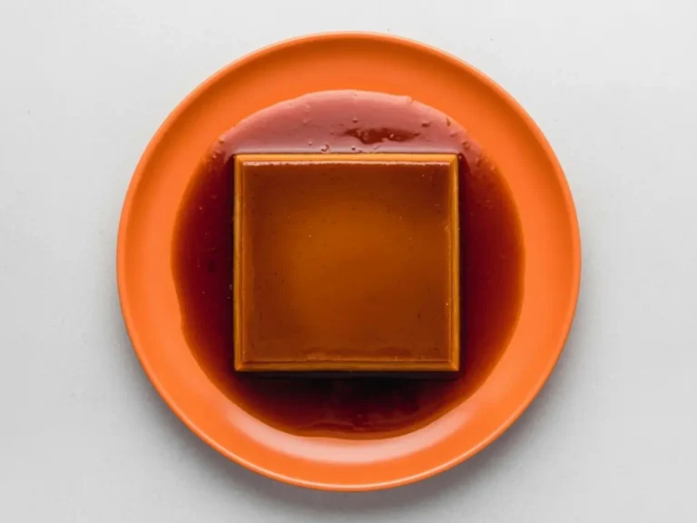 A high-angle 4:3 shot of a rectangular Dulce de Leche Crema Volteada (Flan) centered on a minimalist white ceramic plate with a subtle honeycomb texture. The flan is a rich golden-tan color, sitting in a generous pool of dark amber translucent caramel syrup. The scene is set on a white marble countertop with soft morning light from the side. In the background, a small wooden cutting board and a tiny sprig of mint are softly blurred. Professional food photography style, warm tones, clean and elegant.