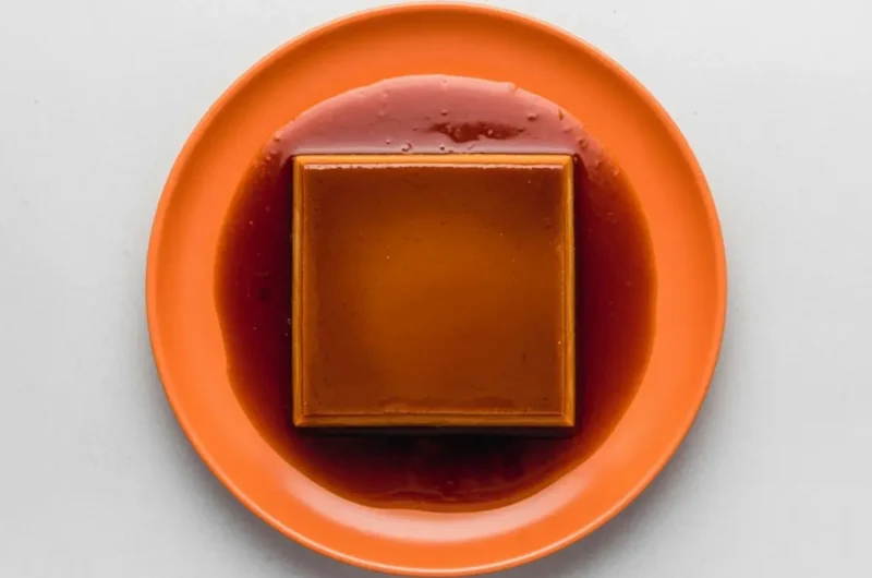 A high-angle 4:3 shot of a rectangular Dulce de Leche Crema Volteada (Flan) centered on a minimalist white ceramic plate with a subtle honeycomb texture. The flan is a rich golden-tan color, sitting in a generous pool of dark amber translucent caramel syrup. The scene is set on a white marble countertop with soft morning light from the side. In the background, a small wooden cutting board and a tiny sprig of mint are softly blurred. Professional food photography style, warm tones, clean and elegant.