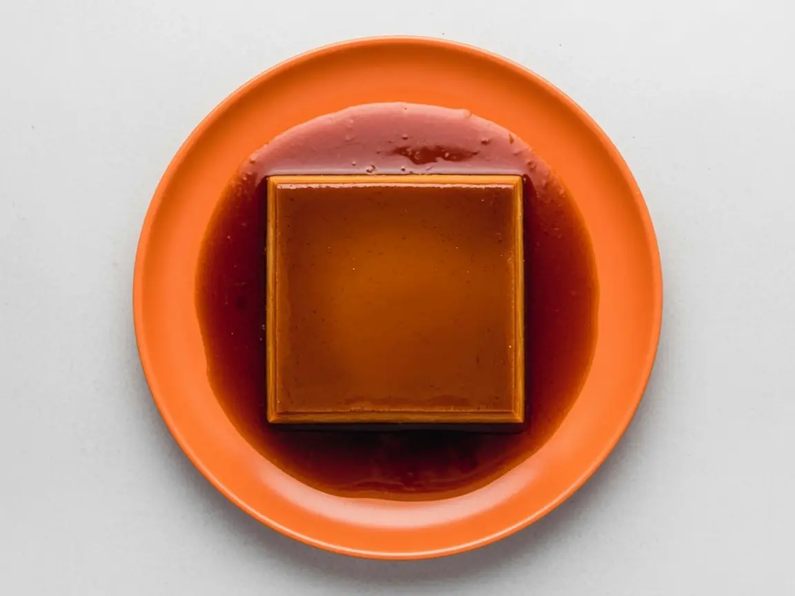 A high-angle 4:3 shot of a rectangular Dulce de Leche Crema Volteada (Flan) centered on a minimalist white ceramic plate with a subtle honeycomb texture. The flan is a rich golden-tan color, sitting in a generous pool of dark amber translucent caramel syrup. The scene is set on a white marble countertop with soft morning light from the side. In the background, a small wooden cutting board and a tiny sprig of mint are softly blurred. Professional food photography style, warm tones, clean and elegant.