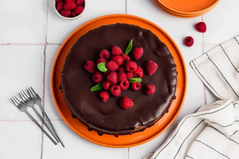A high-angle 4:3 hero shot of a whole Triple Chocolate Cheesecake with Chocolate Ganache resting on a white marble slab. The cheesecake has a dark, thick chocolate cookie crust, a rich chocolate filling, and a glossy dark chocolate ganache dripping down the sides. A pile of vibrant red raspberries and a small mint sprig sit in the center. The background is a clean white tiled wall with soft morning light from an east window, creating gentle shadows. A striped linen napkin is tucked partially under the marble slab.