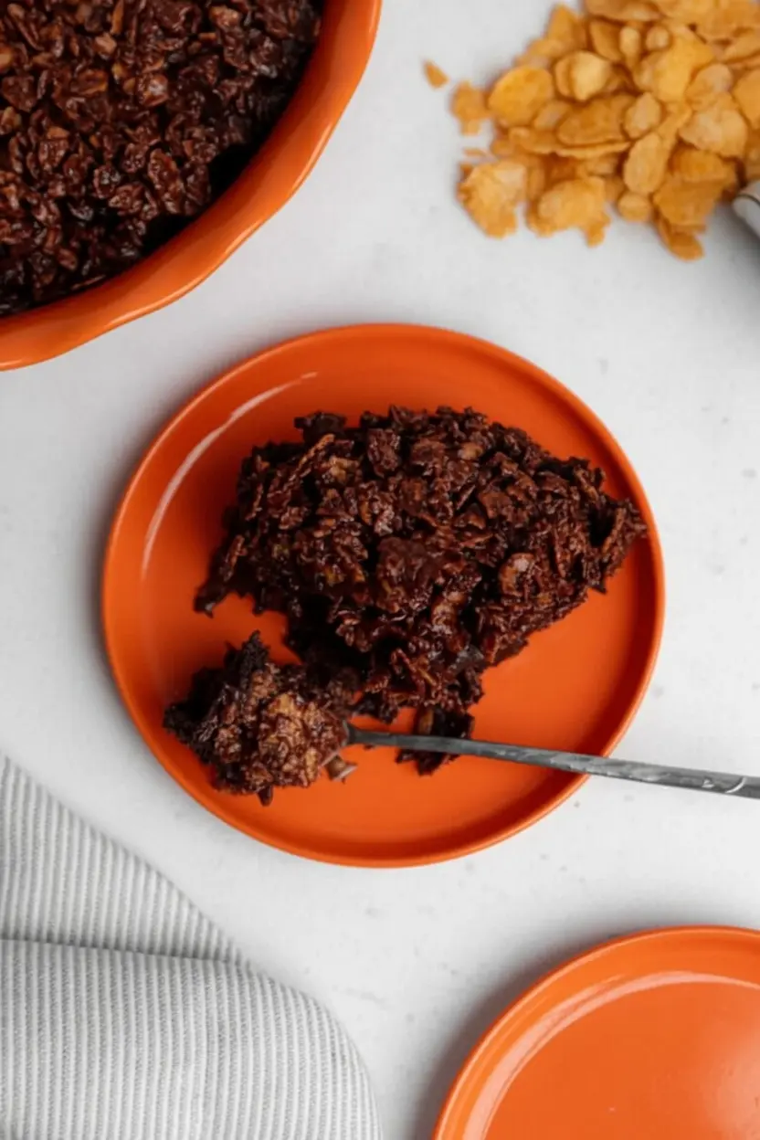 A process shot showing a wooden spoon stirring thick, glossy melted dark chocolate into a pile of cornflakes in a white ceramic bowl. The chocolate is coating the flakes, creating a rich, textured mixture. The scene is clean and tidy with a wooden cutting board in the background.