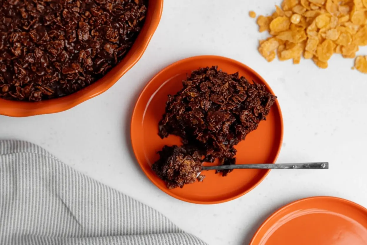 A hero shot of a thick square of Chocolate Crunch Cake on a minimalist white ceramic plate. The cake has a dark, glossy chocolate finish with highly visible, jagged textures of chocolate-coated cornflakes. A silver fork rests on the plate with a small bite taken out, revealing the fudgy interior. The scene is set on a white marble countertop with soft morning light from an east window casting gentle shadows. Fresh mint is visible in the soft-focus background.